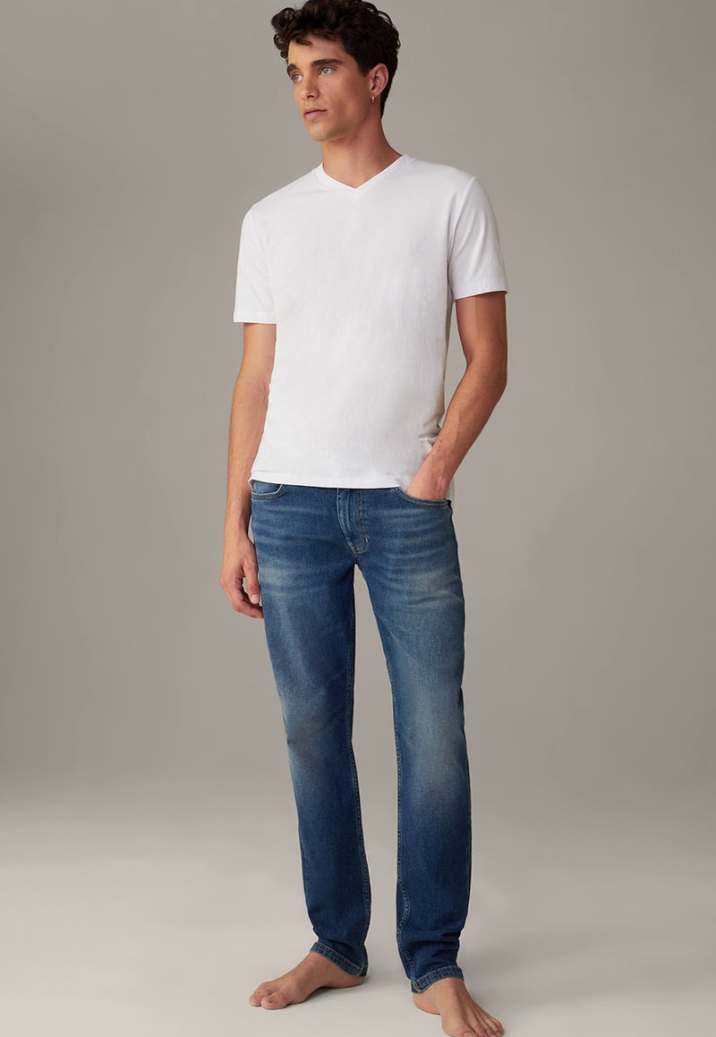 Weißes Baumwoll-V-Ausschnitt-T-Shirt kombiniert mit blauen Jeans, das eine lockere Passform und sanfte Verblassung im Stoff aufweist. Barfußstellung.