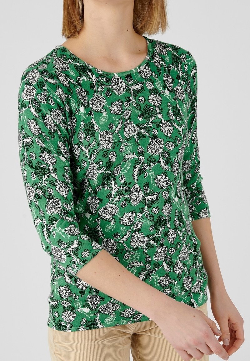 Femme portant une chemise verte à manches longues avec un motif floral blanc et noir et un pantalon beige sur un fond uni.