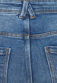 Tela de mezclilla azul con costuras naranjas que muestra los lazos traseros del cinturón y los bordes superiores de dos bolsillos traseros.