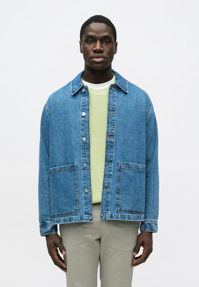 Jeune homme portant une veste en denim bleue avec des poches avant, par-dessus un pull vert clair et un pantalon beige, debout devant un fond blanc uni.
