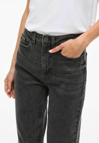 Crne traperice s uskim fitom, visokim strukom i prednjim džepovima. Sadrže metalnu kopču i petlje za remen. Tkanina pokazuje lagane varijacije u teksturi.