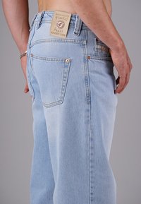 Mann trägt hellblaue Jeans mit sichtbaren Gesäßtaschen und einem hellbraunen Lederpatch mit der Aufschrift "Picaldi" am Bund.
