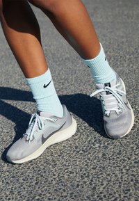 Scarpe da ginnastica grigie con dettagli azzurri e lacci bianchi. Indossate con calze Nike azzurre chiare con un logo nero. Texture del terreno grezzo visibile.