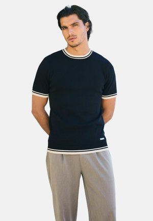 Pull à manches courtes bleu marine avec col et poignets côtelés couleur crème, associé à un pantalon gris clair. Texture lisse, coupe ajustée.