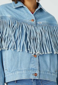 Světle modrá džínová bunda s třásněmi na přední straně, měděnými knoflíky a krátkým střihem se střihnutým lemem.