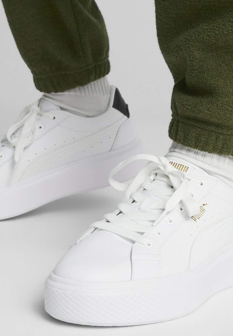 Białe skórzane sneakersy z wzorzystymi teksturowanymi bokami, zaokrąglonymi czubkami i grubą białą podeszwą. Czarne akcenty na pięcie. Sznurówki i logo w kolorze złotym.