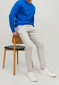 Lysegrå, tætsiddende bukser, blå sweatshirt med rund hals og hvide sneakers. Modellen sidder på en træstol med sort sæde.