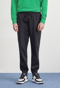 Pantaloni della tuta neri con vita elastica e coulisse regolabile, abbinati a sneaker bianche e nere. Felpa verde visibile nella parte superiore.