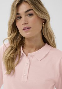 Polo rose clair en tissu texturé, avec col classique et patte à trois boutons. Le tissu semble doux et ajusté.