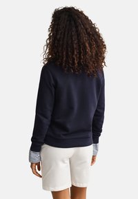 Person med krøllete hår som har på seg en marineblå genser over en stripete langermet skjorte og hvite shorts, vendt bort fra betrakteren.