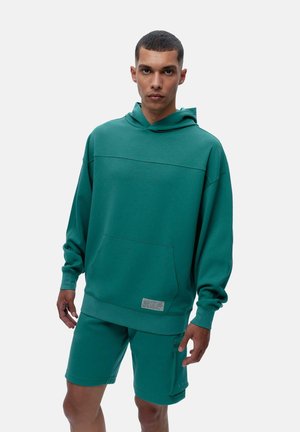Teal hoodie med en framficka och subtila tonala sömmar, matchat med shorts som har en cargoficka och tryckknappsdetaljer.