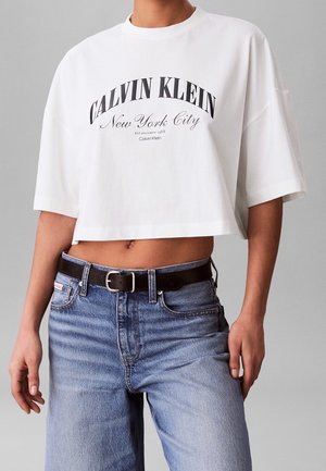 T-shirt court blanc Calvin Klein avec texte noir "Calvin Klein New York City", porté avec un jean bleu et une ceinture noire.
