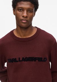 Maglione bordeaux con texture a maglia, caratterizzato da un logo ricamato nero "KARL LAGERFELD" ben visibile sul petto e un colletto a coste.