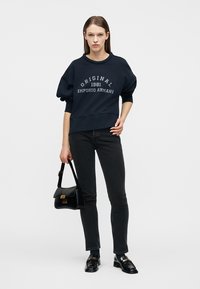 Νέα γυναίκα με μαύρο φούτερ «Original 1981 Emporio Armani», μαύρο τζιν, μαύρα μοκασίνια, κρατώντας μαύρο τσάντα χειρός.