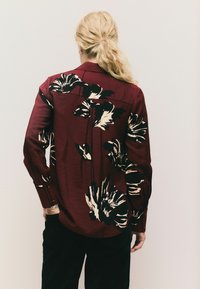 Cămașă roșie cu design floral negru, mâneci lungi și manșete cu nasturi. Țesătură texturată, guler tradițional și detaliu de yoke pe spate.