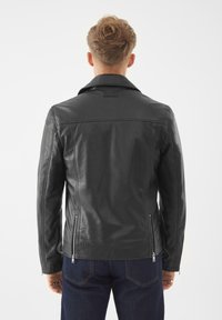 Mann mit kurzem blonden Haar, der eine enganliegende schwarze Lederjacke und dunkelblaue Jeans trägt, von hinten vor einem schlichten weißen Hintergrund gezeigt.