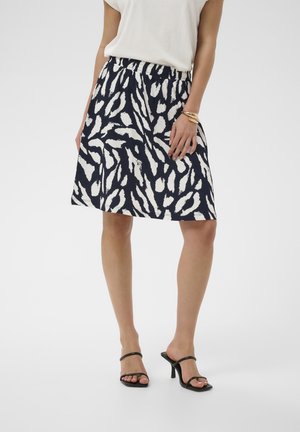 Femme portant une jupe mi-longue noire et blanche à motif abstrait, un chemisier blanc, des sandales noires à talons et des bracelets dorés à un poignet.