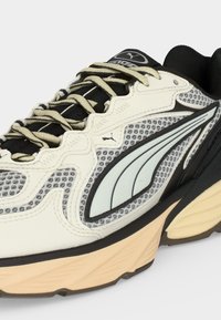 Αθλητικό μαύρο, άσπρο και μπεζ sneaker Puma με λεπτομέρειες από δίχτυ, χοντρή σόλα και χαρακτηριστικό καμπυλωτό λογότυπο στη πλευρά και μπεζ κορδόνια.