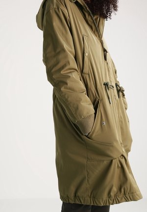 Personne portant un parka à capuche vert olive avec cordons et mains dans les poches latérales, montrée de la moitié du torse vers le bas.
