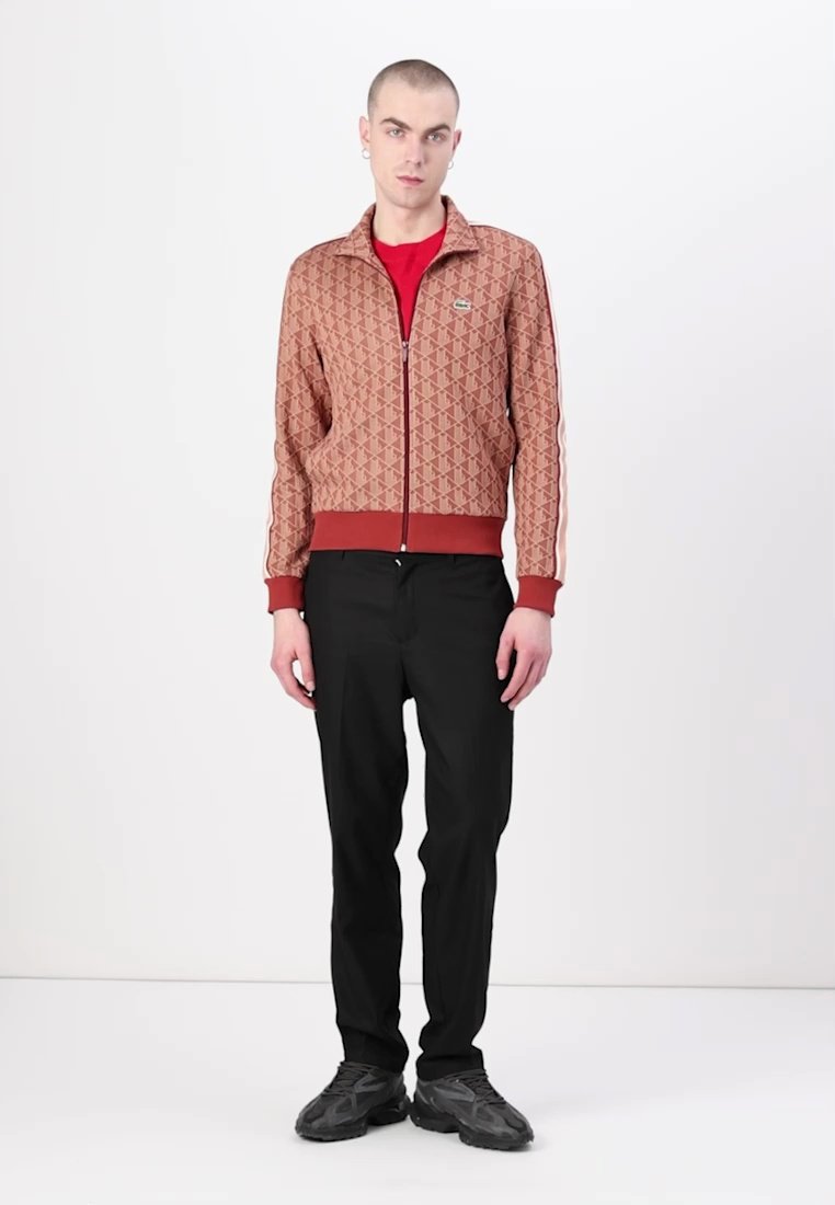 Veste en cuir marron avec un motif géométrique, col et poignets côtelés rouges, assortie à un pantalon noir et des chaussures grises. Le mannequin se tient debout.
