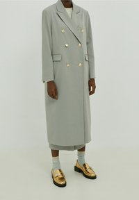 Manteau gris croisé à boutons dorés, avec col cranté et poches latérales. Associé à des mocassins dorés brillants et des chaussettes à rayures.