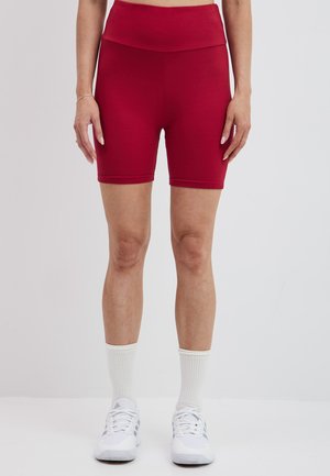 Czerwone rowerowe spodenki o wysokim stanie, wykonane z elastycznego materiału, charakteryzujące się gładką fakturą i dopasowanym krojem, zestawione z białymi trampkami i skarpetkami.