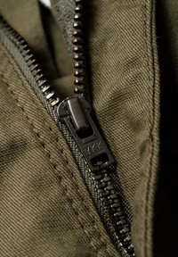 Nahaufnahme von olivgrünem Stoff mit einem schwarzen Metallreißverschluss mit YKK-Branding, der die strukturierte Webart und die Nähdetails hervorhebt.