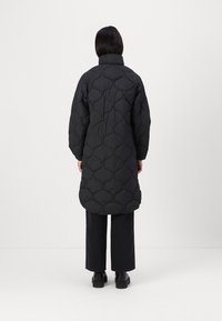 Svart quiltad lång kappa med hög krage, med rundade sömmönster. Bärs över svarta vida byxor och kraftiga svarta stövlar.