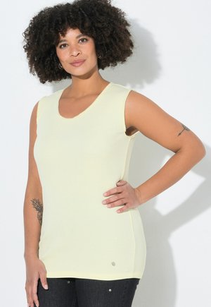 Mujer con cabello rizado, vestida con una blusa sin mangas de color amarillo claro y pantalones negros, parada con una mano en la cadera frente a un fondo blanco.