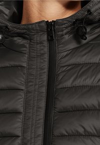 Czarna kurtka puchowa z teksturowanym, pikowanym wzorem, z przednim zamkiem błyskawicznym i regulowanymi sznurkami w kapturze. Miękki, błyszczący materiał.