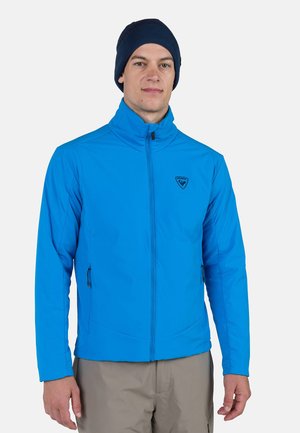 Leichte blaue Jacke mit hohem Kragen, durchgehendem Reißverschluss und seitlichen Taschen. Mit einem Logo auf der Brust und kombiniert mit einer marineblauen Mütze.