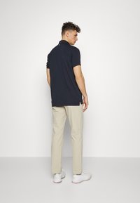 Camisa polo azul marino de manga corta con un ligero dobladillo asimétrico, combinada con pantalones beige y zapatillas deportivas blancas, vista desde la parte de atrás.