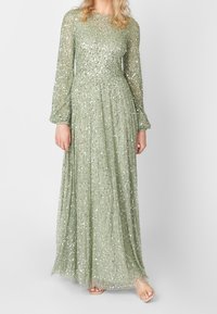 Lång, ljusgrön klänning med genomskinliga långärmar, dekorerad med spridda paljetter, med en flödande design och veckad struktur.