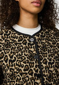 Leopardmönstrad jacka i beige och svart, med rund halsringning, svarta detaljer och knappar framtill. Mjuk tyg med en texturerad yta.