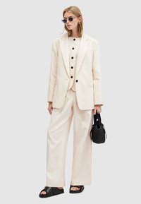 Abito beige a righe sottili con giacca su misura e pantaloni a gamba larga. Bottoni neri e una borsa nera texturizzata. Occhiali da sole a forma di occhio di gatto di grandi dimensioni.