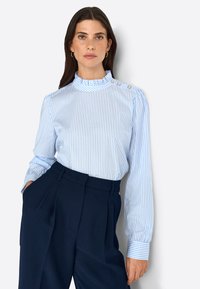 Blusa a rayas azul claro con un alto cuello con volantes y detalles de botones. Mangas largas y tejido suave. Combinada con pantalones oscuros de navy.