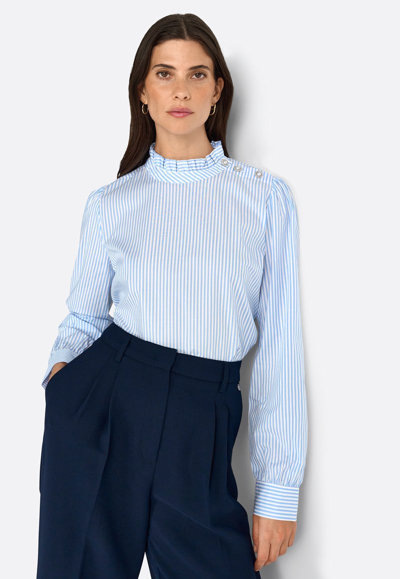 Blusa a rayas azul claro con un alto cuello con volantes y detalles de botones. Mangas largas y tejido suave. Combinada con pantalones oscuros de navy.