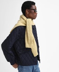 Navy quilted jacket with large buttons, paired with a yellow cable-knit scarf draped over the shoulders. Model wears black sunglasses.
