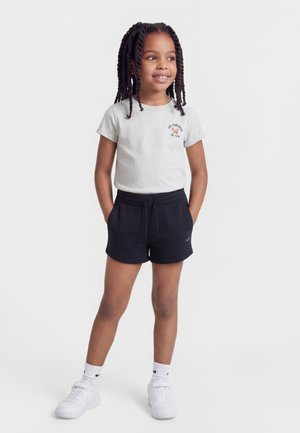 Niña joven con cabello trenzado, vestida con una camiseta gráfica blanca, pantalones cortos negros, calcetines blancos y zapatillas blancas, de pie con las manos en los bolsillos.