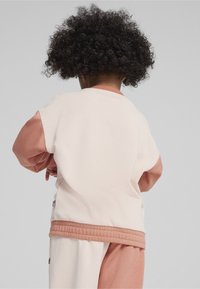 Ljus krämfärgad sweatshirt med rosa accenter, ribbat nederkant och ärmslut. Baksidan har en slät textur och en avslappnad passform, buren av ett barn.