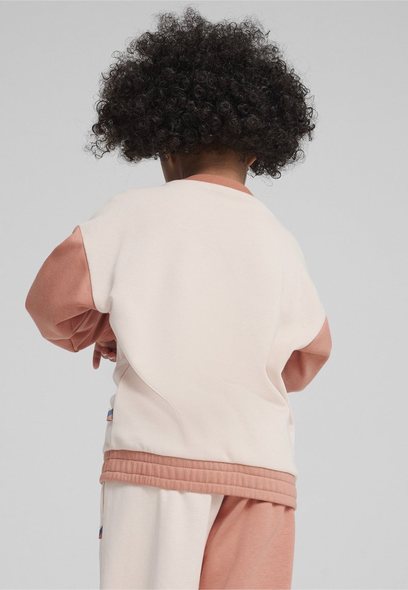 Ljus krämfärgad sweatshirt med rosa accenter, ribbat nederkant och ärmslut. Baksidan har en slät textur och en avslappnad passform, buren av ett barn.