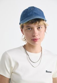 Cappello blu di jeans con visiera curva e dettagli di cucitura, abbinato a una maglietta bianca a coste con un piccolo logo. Due collane d'argento sovrapposte.