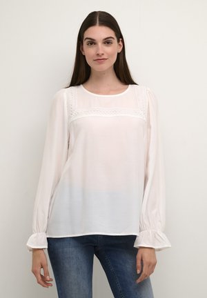 Vit långärmad blus i lätt tyg, med spetsdetalj över bröstet och volangärmar. Modellen har på sig blå jeans.