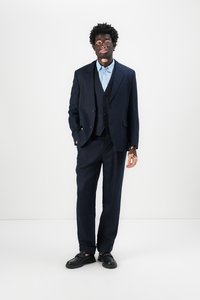 Maßgeschneideter marineblauer Anzug mit einer Jacke mit drei Knöpfen, passender Weste und schmal geschnittenen Hosen, kombiniert mit einem hellblauen Hemd und schwarzen Slippern.