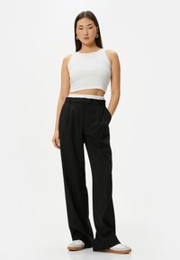Zwarte wijde broek met een ceintuur in de taille, gecombineerd met een cropped witte tanktop. Het model draagt witte schoenen met een beige zool.