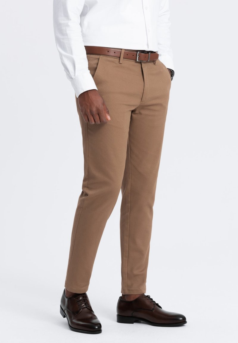 Pantaloni chinos marroni con una finitura testurizzata e una vestibilità su misura, abbinati a una cintura in pelle marrone e scarpe eleganti marroni scuri. Camicia bianca a maniche lunghe.