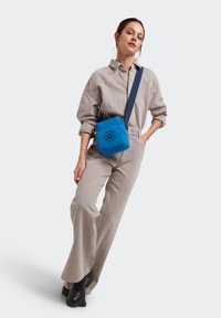 Blå, kviltad axelväska med fyrkantig form och en rund logotyp på framsidan. Modellen bär en ljusgrå jumpsuit och svarta stövlar.
