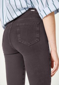 Donkergrijze jeans hebben een slim fit ontwerp met een middelhoge taille, achterzakken en zichtbare stiksels. De textuur lijkt glad en rekbaar.