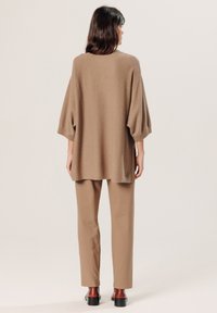 Beige oversized trui met brede mouwen, gecombineerd met lichtbeige rechte broek. Model draagt donkere bruine blokhakken.