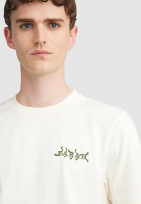 Crème-kleurige katoenen t-shirt met een groen abstract logo op de linkerborst, voorzien van een klassieke ronde hals en korte mouwen.